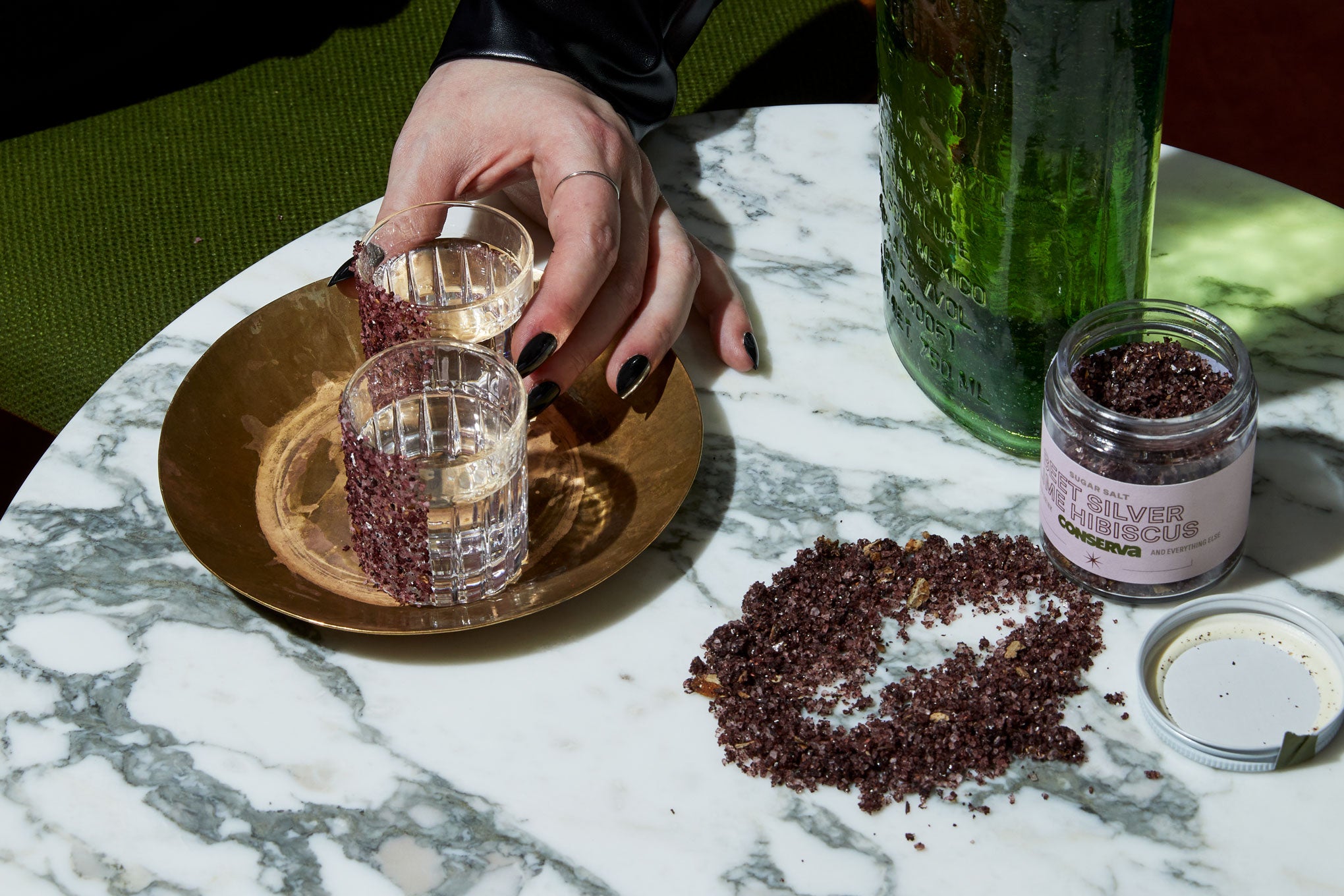 Beet-Hibiscus-Lime Sugar Salt on the side of 2 shot glasses being picked up by a hand on a marble table with tequila