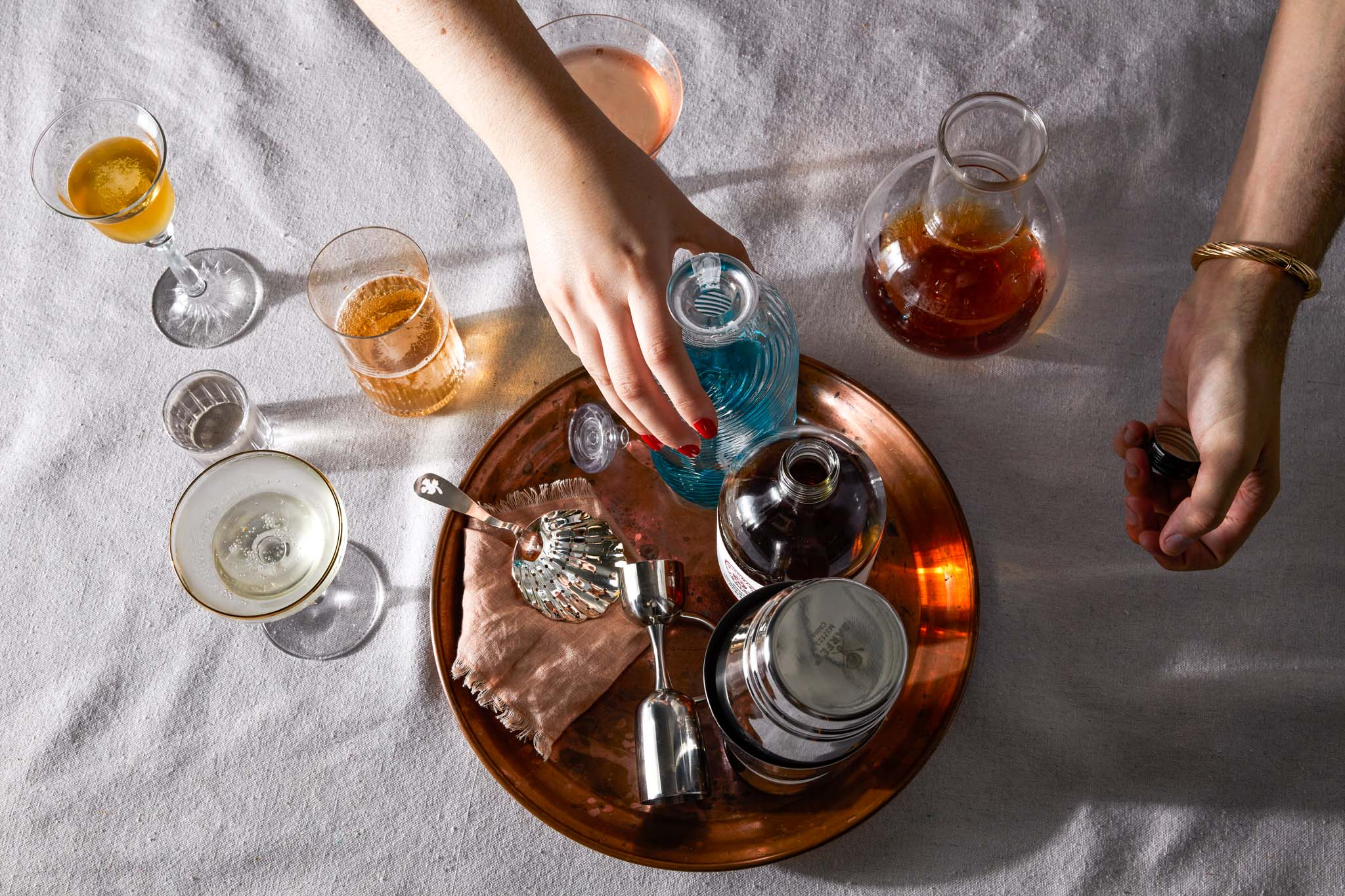 Various cocktail equipment and bottles on a bronze tray, slightly aged, with surrounding glassware on white table cloth. Two hands reach in to grab the bottles. 